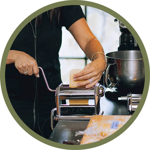 Close-up of hands using a pasta machine to roll fresh dough on a kitchen counter, with cooking utensils in the background.
