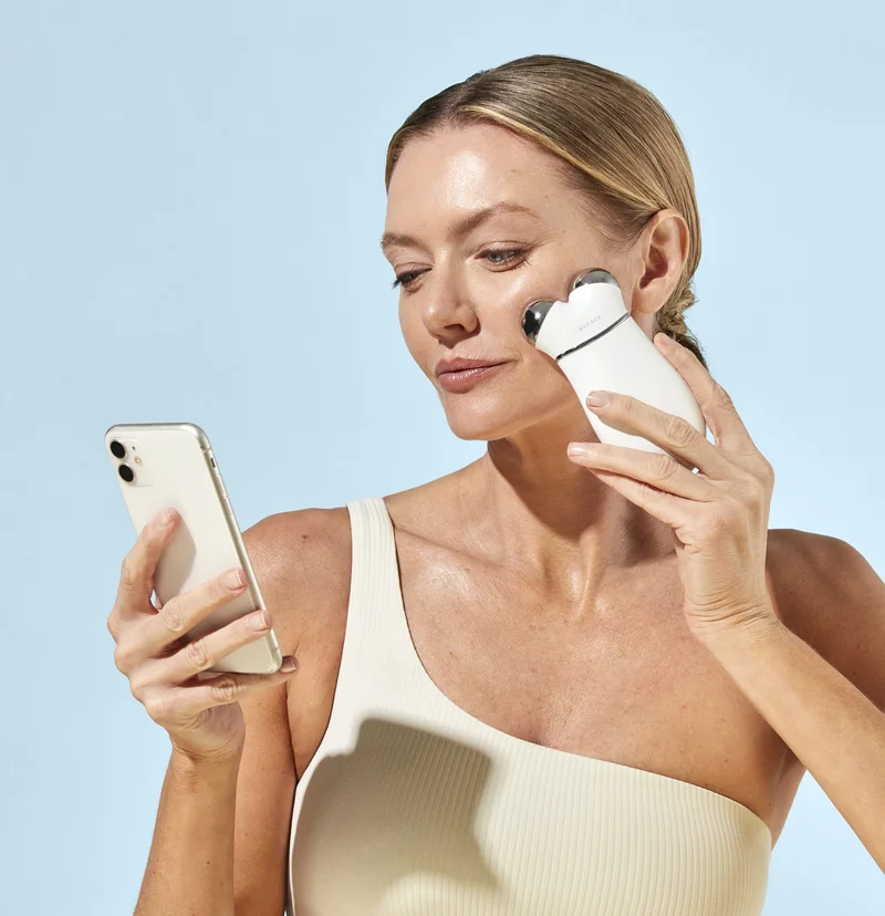 Blonde woman with hair slicked back holding along cheek contour with TRINITY+ Device, following along NuFACE Smart App on phone