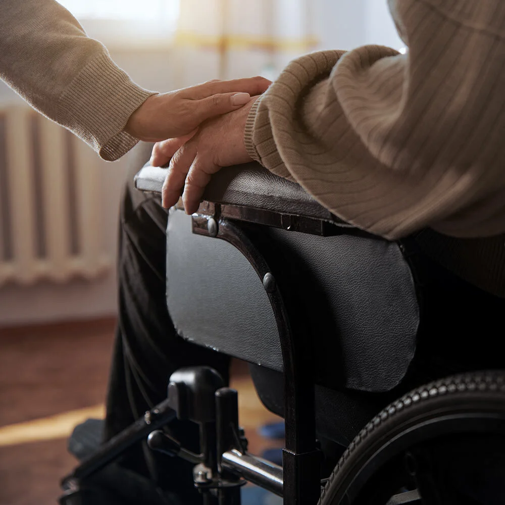 A woman's hand gently resting atop the hand of an older man who is in a wheelchair.