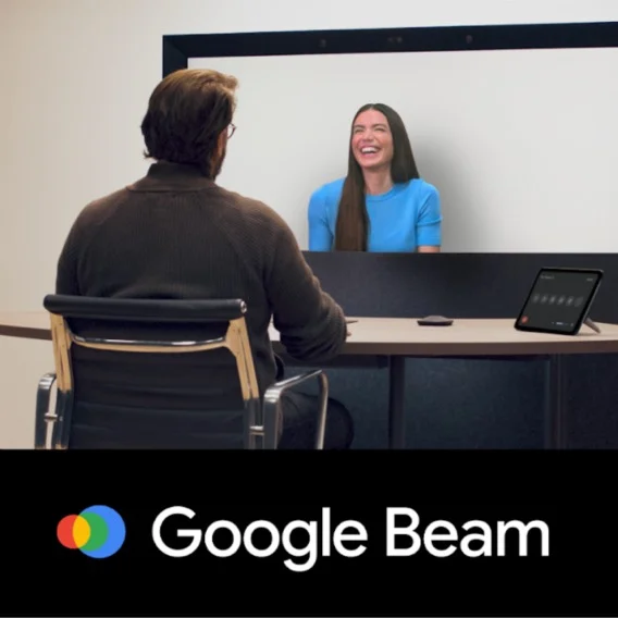 The image shows a 3D video conferencing setup. A man sits at a desk facing a large screen where a woman is displayed. The screen shows Google Beam branding.