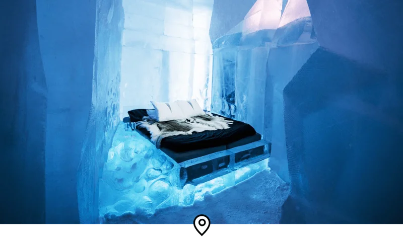 Image Description: Interior of a room in Sweden’s famous Icehotel, featuring a thick mattress with fluffy pillows and reindeer hide on giant ice blocks