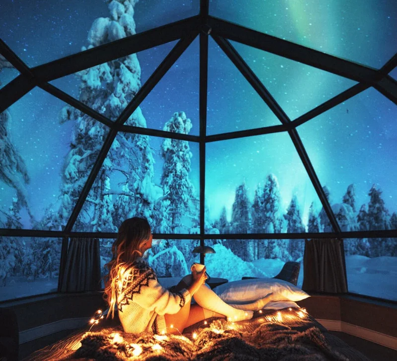 Image Description: A female traveler sitting on a cozy rug in a glass igloo, gazing up at the Northern Lights