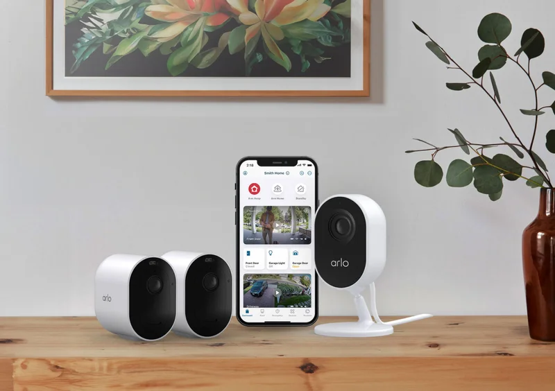 Two Arlo Pro 4 Wireless Security Cameras next to an Essential Wired Indoor Security Camera and a phone displaying a simulated Arlo screen image.