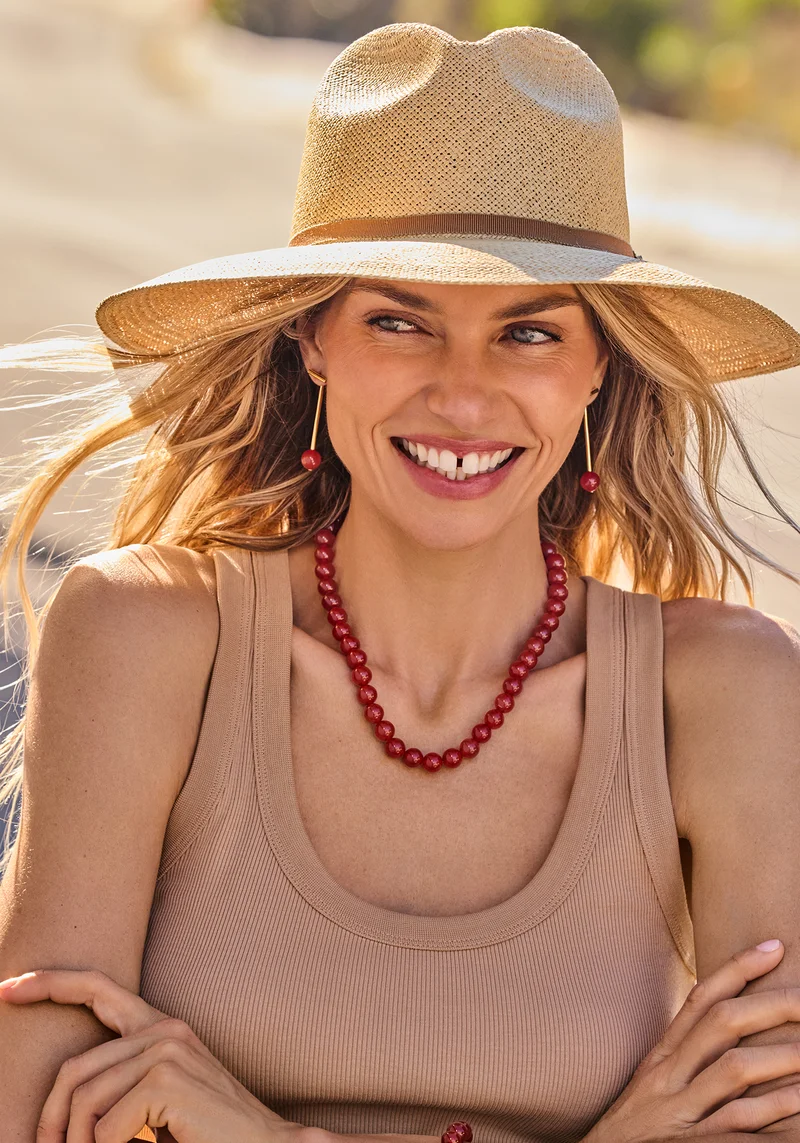 The Fiesta Capsule Packable Wide Brim Ecuador Hat in Dark Natural, Cerise Beaded Necklace in Carnelian Red, Cerise Bar Jacket Earrings in Carnelian Red.