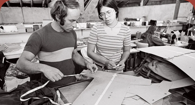 Two people standing at a sewing table taking measurements for a backpack.
