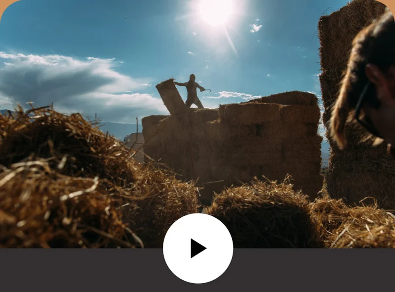 A person stands atop a large stack of hay bales on the back of a flatbed truck.