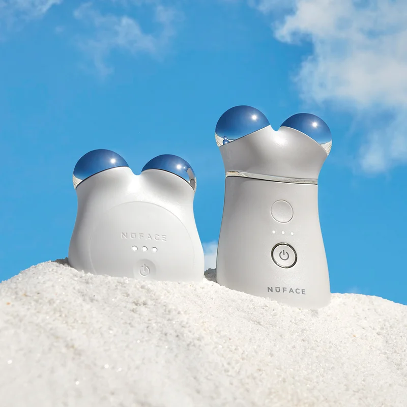NuFACE MINI+ in Brilliant White and TRINITY+ in the sand at the beach against a bright blue sky