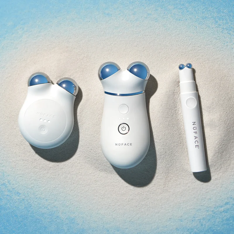 NuFACE MINI+ in Brilliant White, NuFACE TRINITY+, and NuFACE FIX next to each other on a pile of sand against a blue background