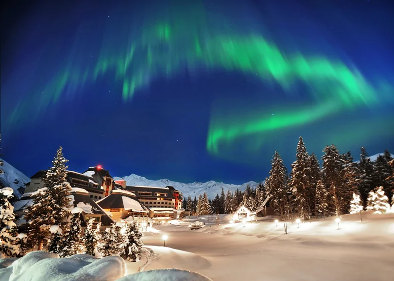 Night view of a ski resort in the snow with colorful swirls of the Northern Lights in the sky.