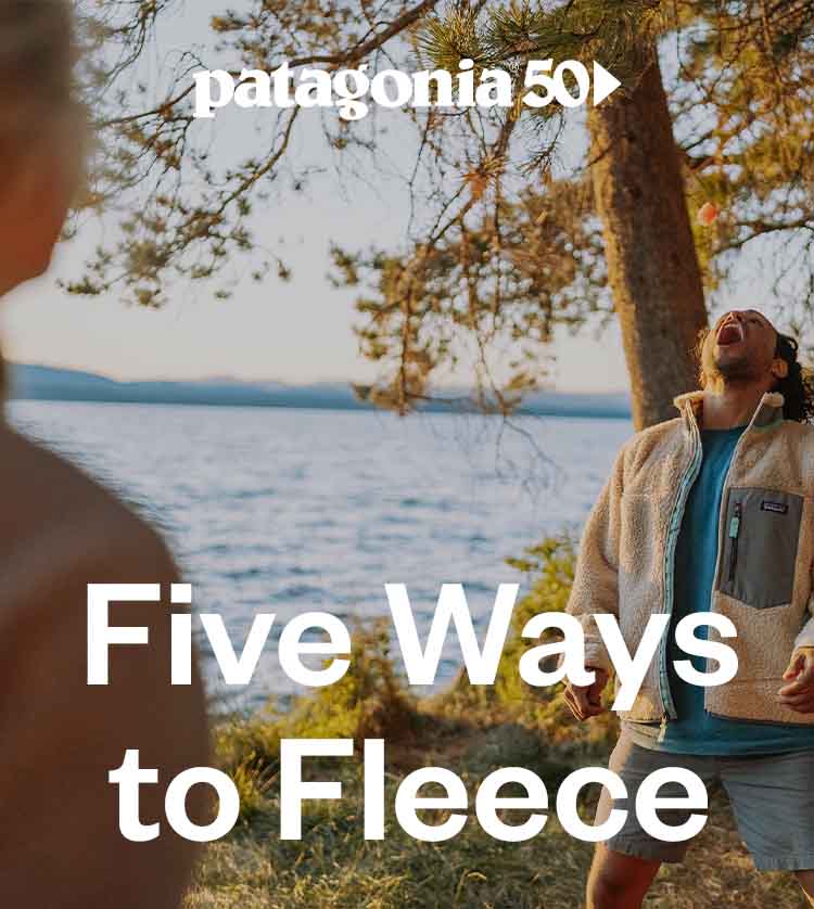 A man attempts to catch a piece of food that a woman tossed to him. He stands on a lake shore surrounded by pine trees with his head tilted back, mouth wide open.