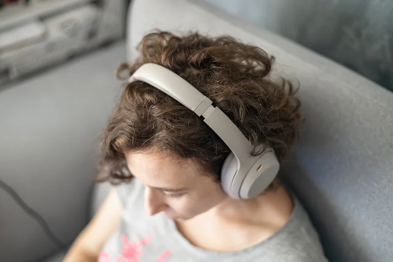 Woman using noise canceling headphones, sitting on gray couch at home.