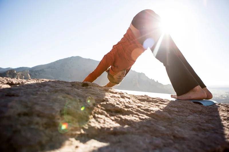 A person practices yoga on a sunny day wearing an orange shirt on top of a big rock with a mountain in the back.