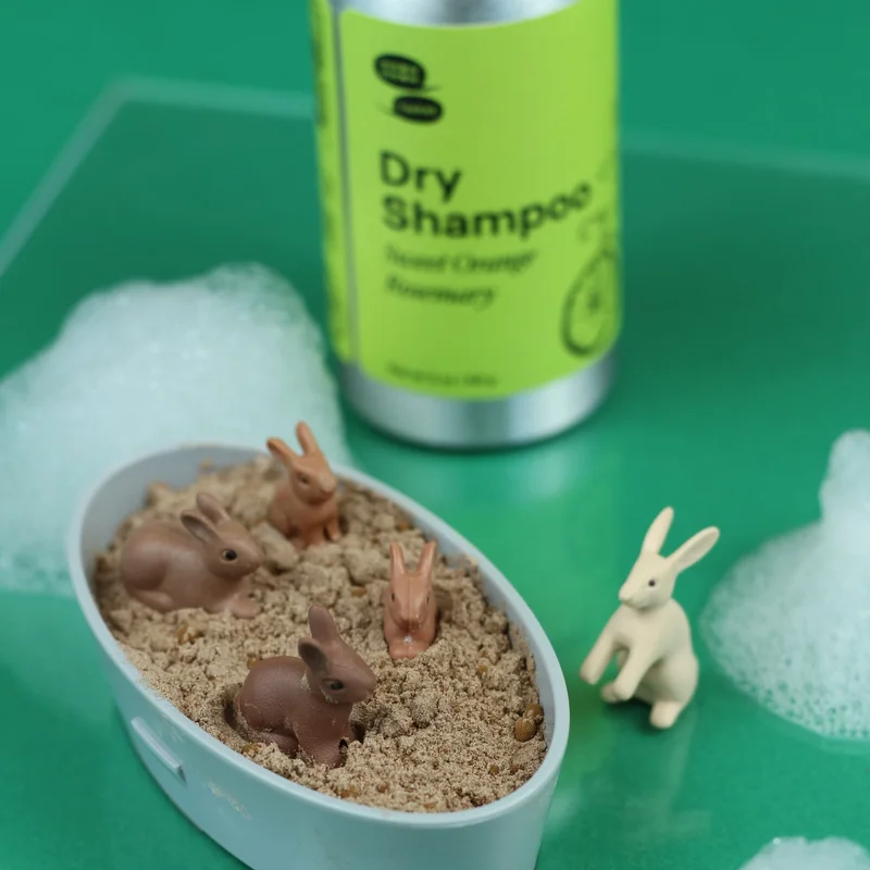 A container filled with shampoo powder has four toy bunnies nestled into the powder with a metal bottle of Dry Shampoo in the background.