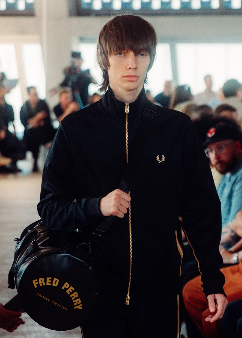 Male model wearing a Fred Perry Black Champagne Track Jacket and carrying a Black / Gold Tonal Barrel Bag