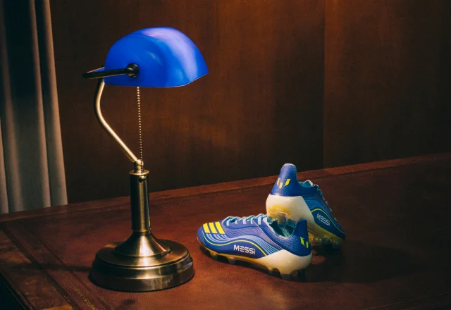The blue and yellow adidas MESSI VIS10N cleats under a blue reading lamp on an antique leather desk.