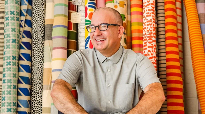 Vince Bravo sits in front of large rolls of colorful patterned textiles by Alexander Girard.
