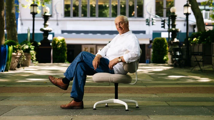 Bruce Hannah sits in a Morrison Hannah Task Chair with beige upholstery and a white base on a sidewalk outside a building in New York