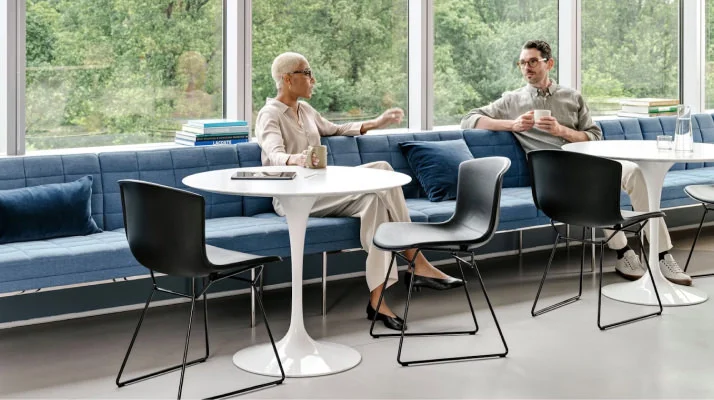 Two individuals conversing in a shared seating area furnished with Saarinen Tables, Knoll Bertoia chairs, and blue bench seating
