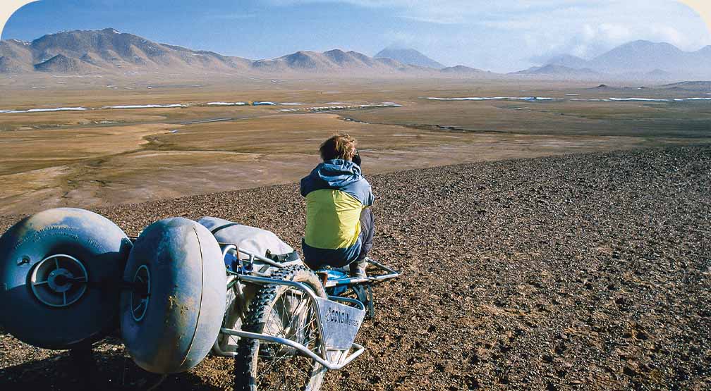 A person sitting on a custom rickshaw loaded with gear looks out over a expansive barren landscape.