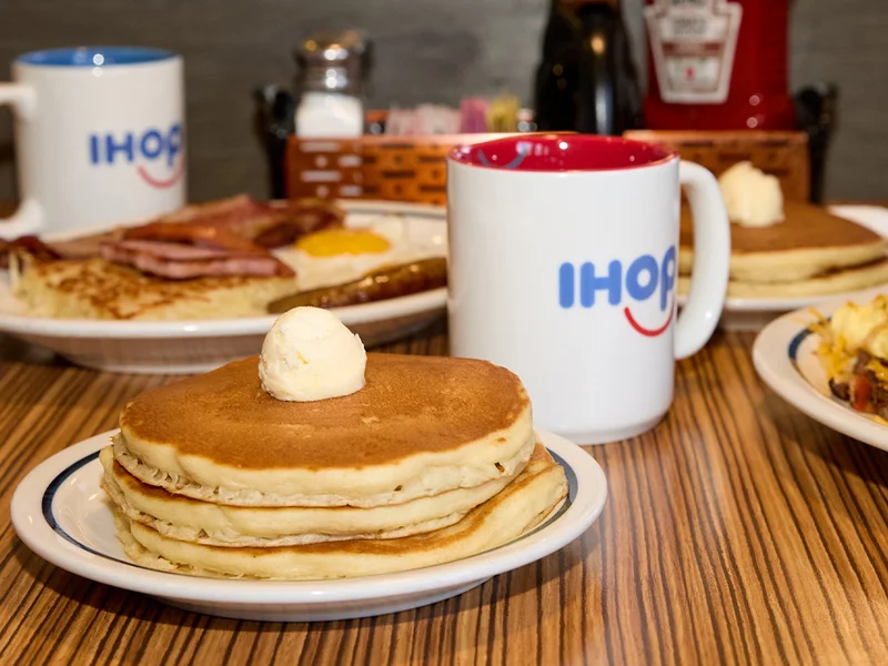 A stack of pancakes topped with butter on a plate, set on a table in a booth at IHOP. In the background is a mug of IHOP coffee and some breakfast meats.