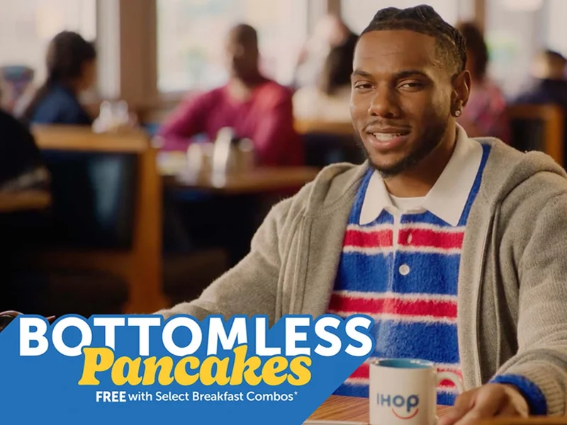 Malik Nabers seated at an IHOP booth, wearing a collared red and blue striped shirt with a grey sweatshirt over it.