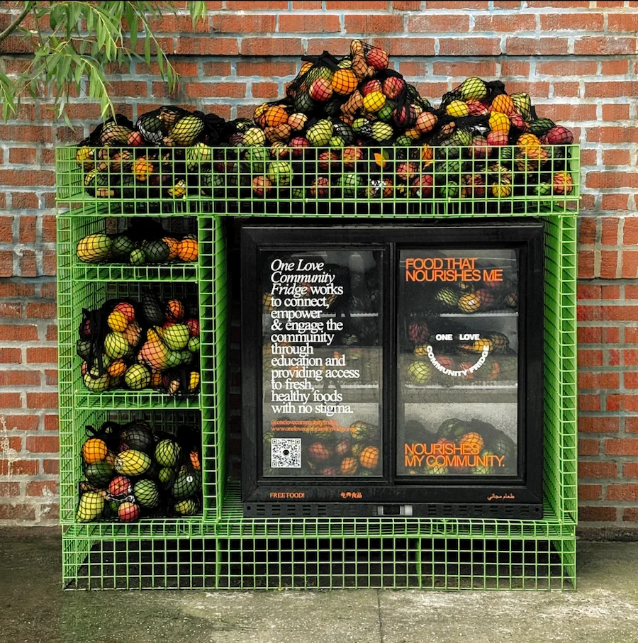 Green apparatus holding bags of fruit, with text reading “One Love Community Fridge works to connect, empower & engage the community through education and providing access to fresh, healthy foods with no stigma.”