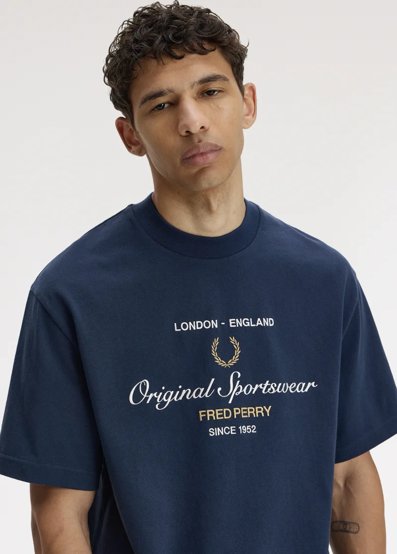 Male model with tanned skin and short curly dark hair looks nonchalantly into the camera and wears an oversized navy T-shirt with white and champagne embroidery at the chest which says "London - England. Original Sportswear, Fred Perry, since 1952".