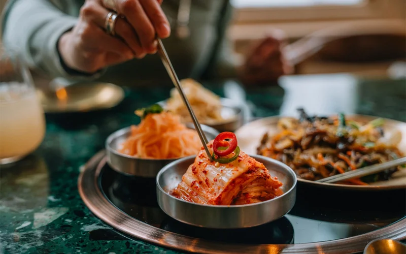 Photo of a hand picking up a piece of kimchi, Korean fermented cabbage, with chopsticks from a table full of assorted food.