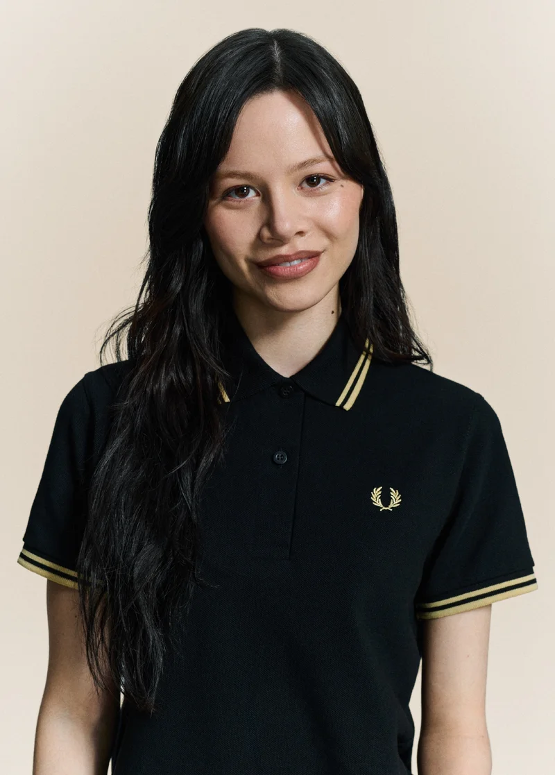 Female model with long black wavy hair smiles and looks directly into the camera. She wears our womenswear version of the Black / Champagne / Champagne Fred Perry Shirt.