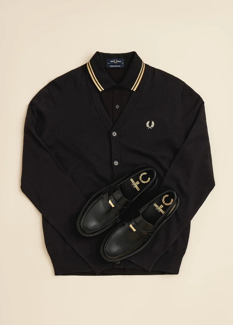 Flat lay image of our black buttoned cardigan, our Black / Champagne / Champagne Fred Perry Shirt and our Fred Perry x George Cox Penny loafers in black