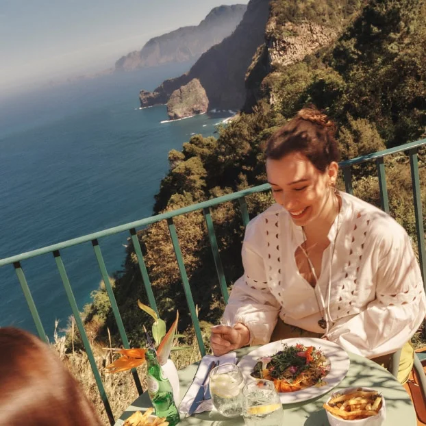 Two people sit opposite each other eating a meal with mountains and the ocean in the background.