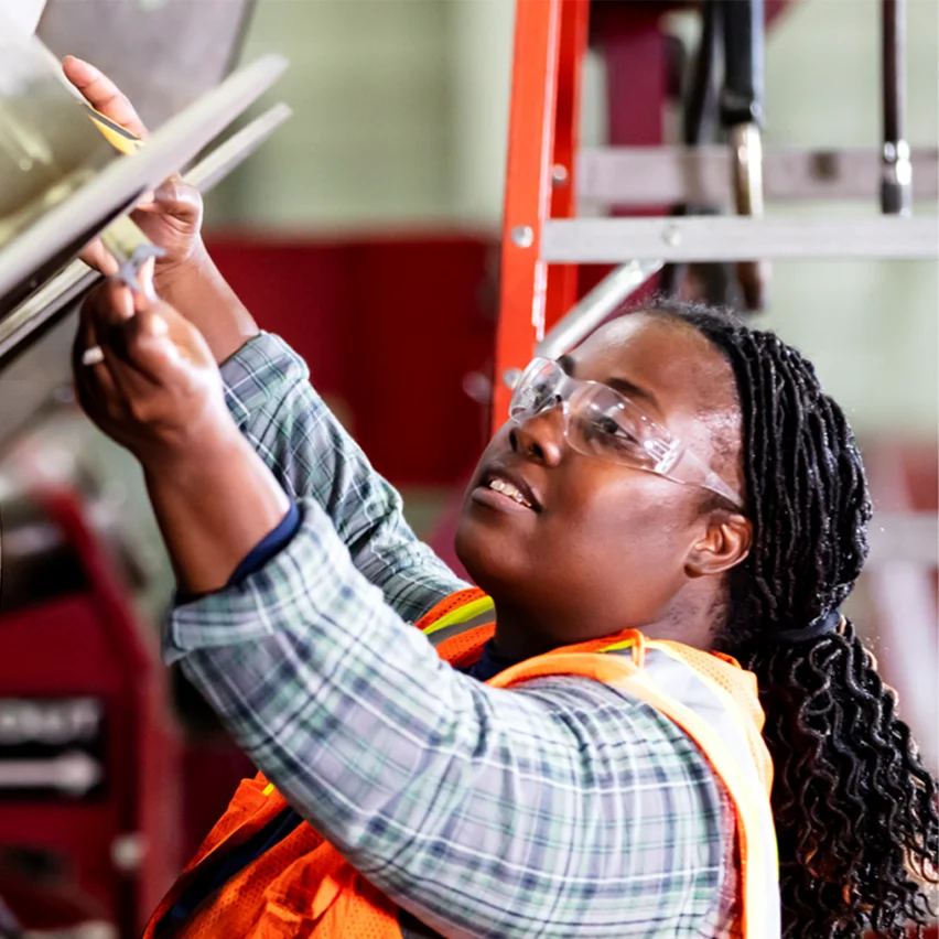 A worker in safety glasses and a high-visibility orange vest uses a measuring tool to inspect a large metal component in an industrial setting.