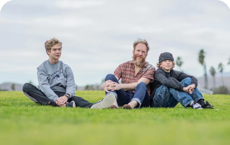 A father sitting in the grass with his two teenage boys.
