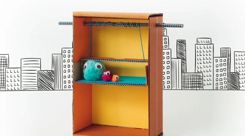 A cardboard elevator lifts a stuffed animal using a string pulley system in front of a cityscape backdrop.