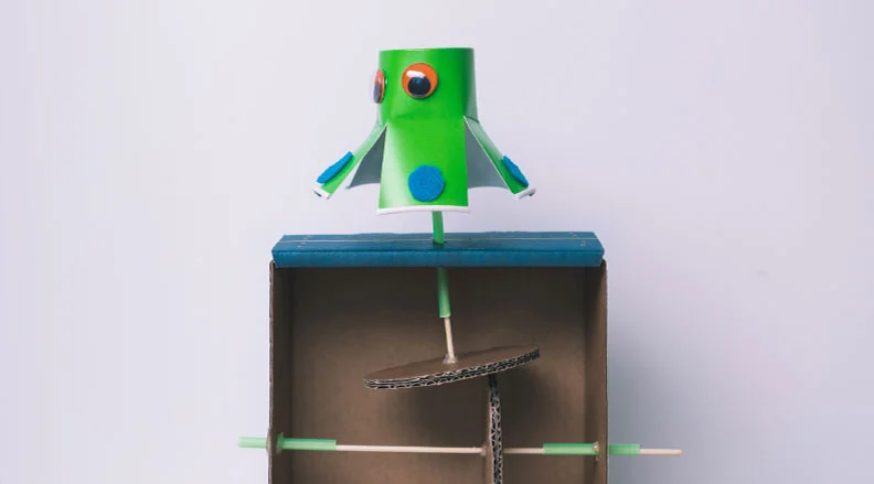 A colorful cardboard robot automaton spins and bounces with personality using a crank system.