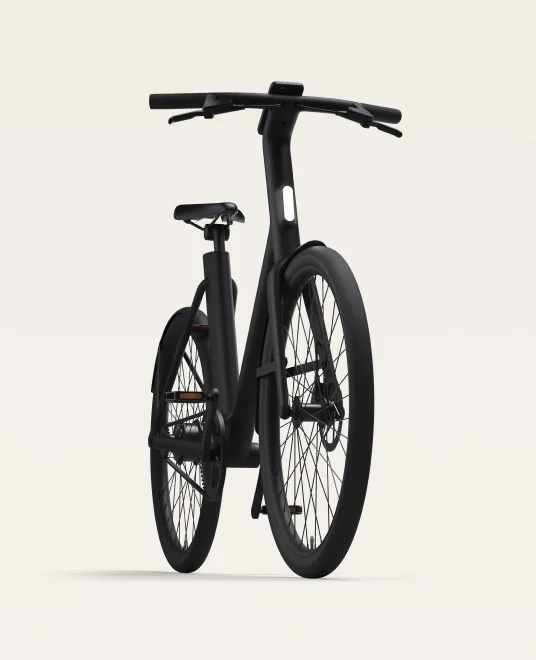 A render. A front view of a black Cowboy e-bike, showcasing its minimalist design with integrated front light, wide handlebars, and sleek frame geometry.