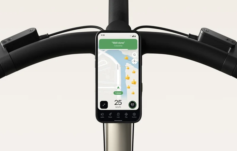 A render. A smartphone mounted on the handlebars of a Cowboy e-bike, displaying a navigation screen with a “Well done” message and a speed of 25 km/h. The app interface shows route details and icons for various bike controls at the bottom.