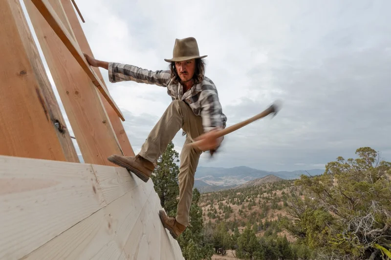 A person hangs off the roof of an unfinished cabin and hammers in a bad nail.