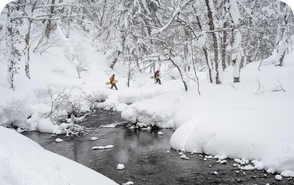 In a forest whitened with fresh snow, two snowboarders walk over a dark river while carrying their boards.