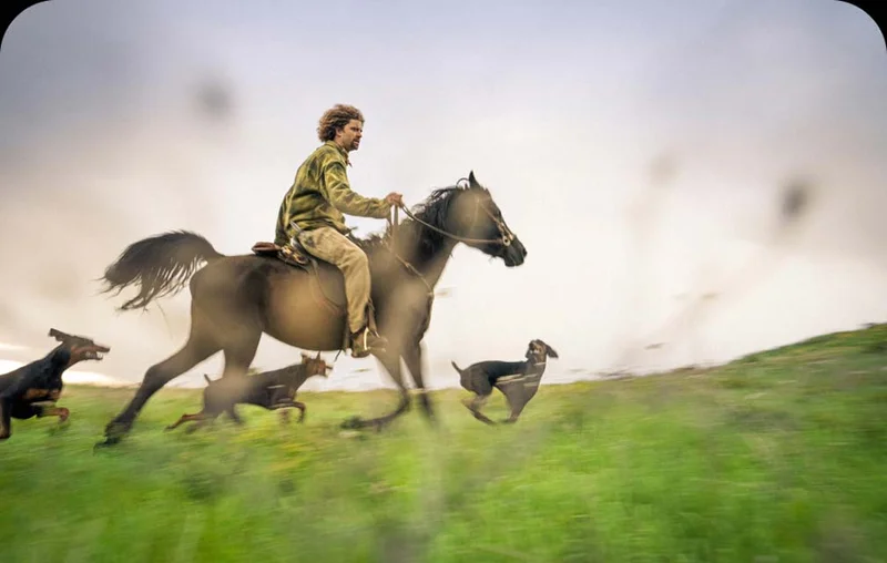 A man rides a galloping horse while his pack of dogs runs alongside.