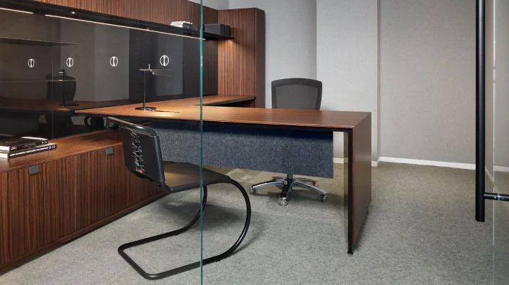 A private office setting with a medium brown wood credenza, height-adjustable desk, wall panel, and overhead from the Knoll Cove Collection. A black side chair and work chair are seated at the desk opposite each other.