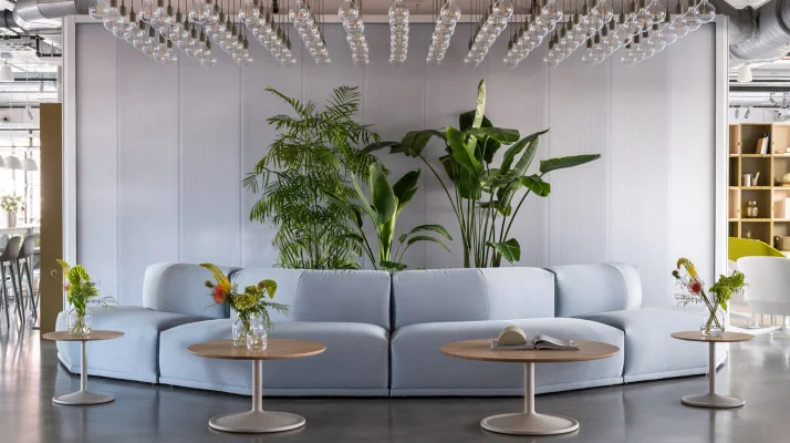 A welcoming lobby lounge with Muuto furniture including a light blue sectional curved around tall green plants with a white wall to the rear, four coffee tables with light wood tops and cream pedestal legs, and rows of white pendant lights hanging above.