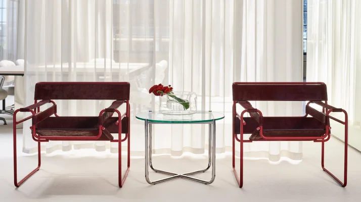 Two dark red modern side chairs seated next to each other, a glass side table with a vase of red flowers on top in between the chairs, and a sheer white curtain in the background.