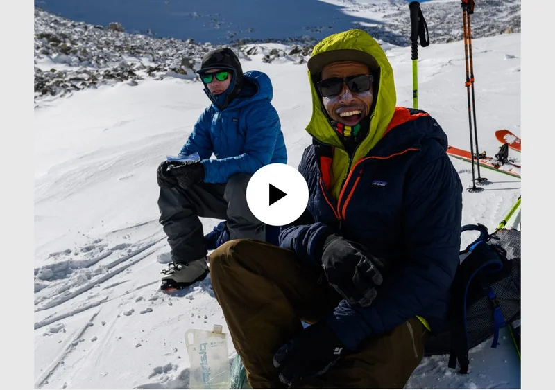 Video play button. Two backcountry skiers sitting on the snow.