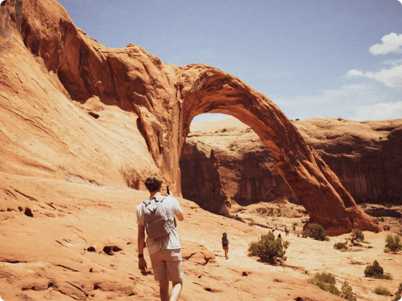 Image Description: Rear view of a traveler hiking towards the Corona Arch, a famous red rock formation in Moab, Utah