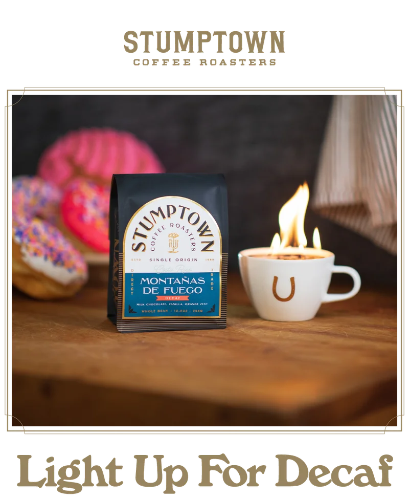 Stumptown single origin Montaña de Fuego decaf coffee bag displayed on a wooden table beside a white coffee cup with a horseshoe logo and flames rising from the cup. Colorful sprinkled donuts sit blurred in the background. Headline reads: “Light Up For Decaf.”