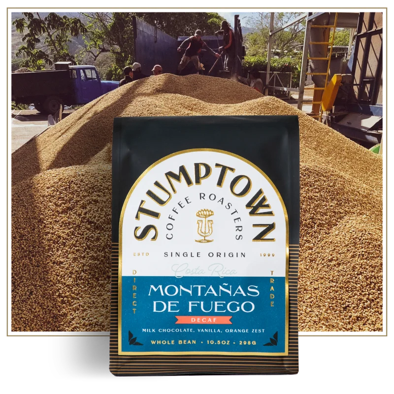 A large mound of drying coffee beans fills the foreground at a Costa Rican coffee mill. A Stumptown Coffee Roasters Montañas de Fuego decaf coffee bag is overlaid in front, highlighting the coffee’s origin and production process.