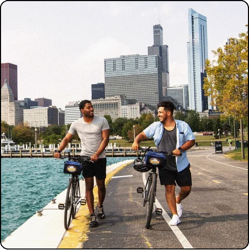 Image Description: Two men smile and look at each other while pushing their bikes on a city bike path along the water.