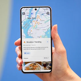 A hand holding a Pixel phone with Google Maps pulled up on the screen. There's a text box that reads 'Brooklyn: Trending' with photos of food beneath.