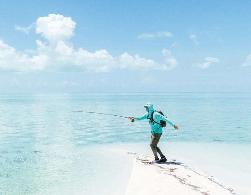 A person stands on white sand and casts a fly rod into tropical waters.